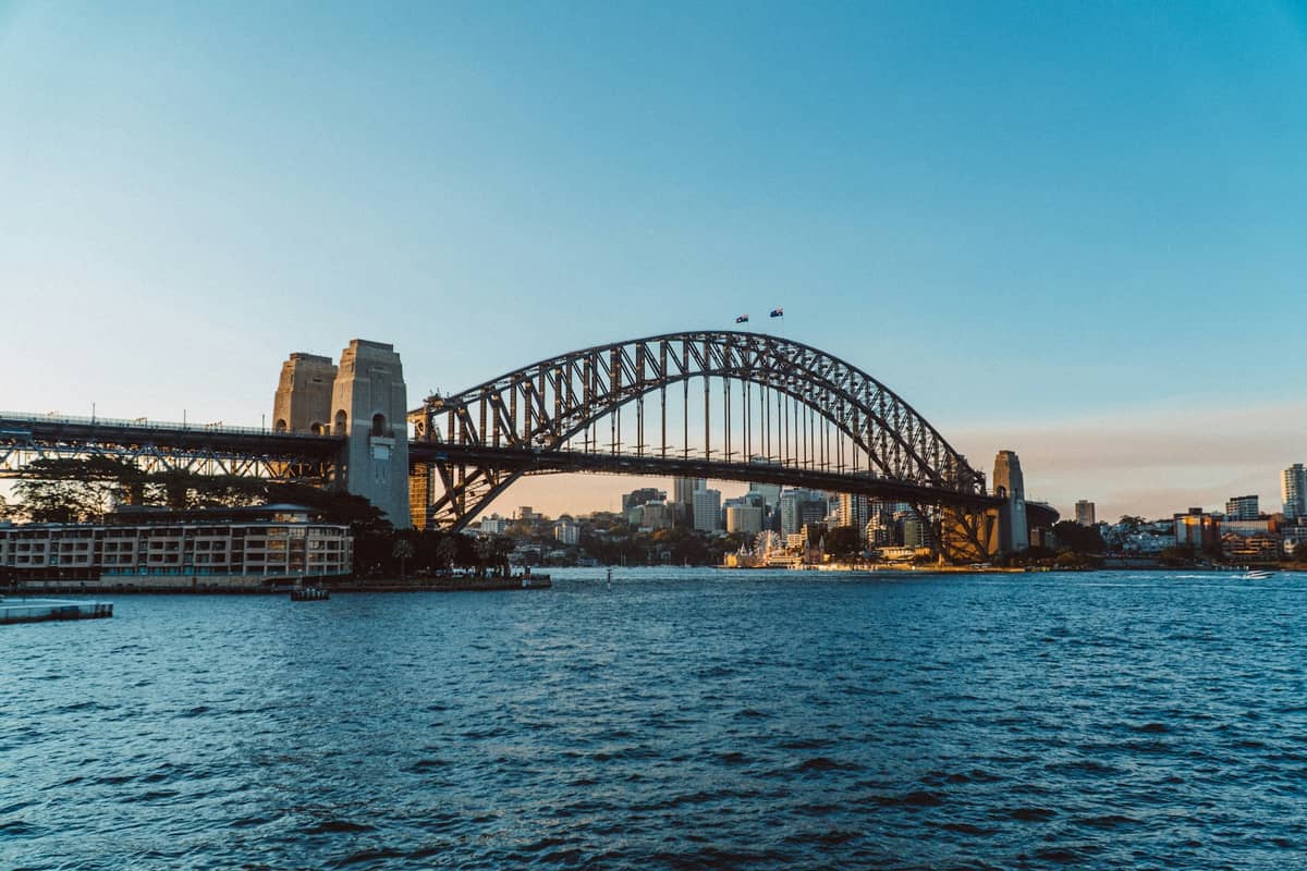 Szeroki widok w ciągu dnia na Most Harbour Bridge przerzucony nad zatoką, z zabudową miejską w tle i bezchmurnym błękitnym niebem.