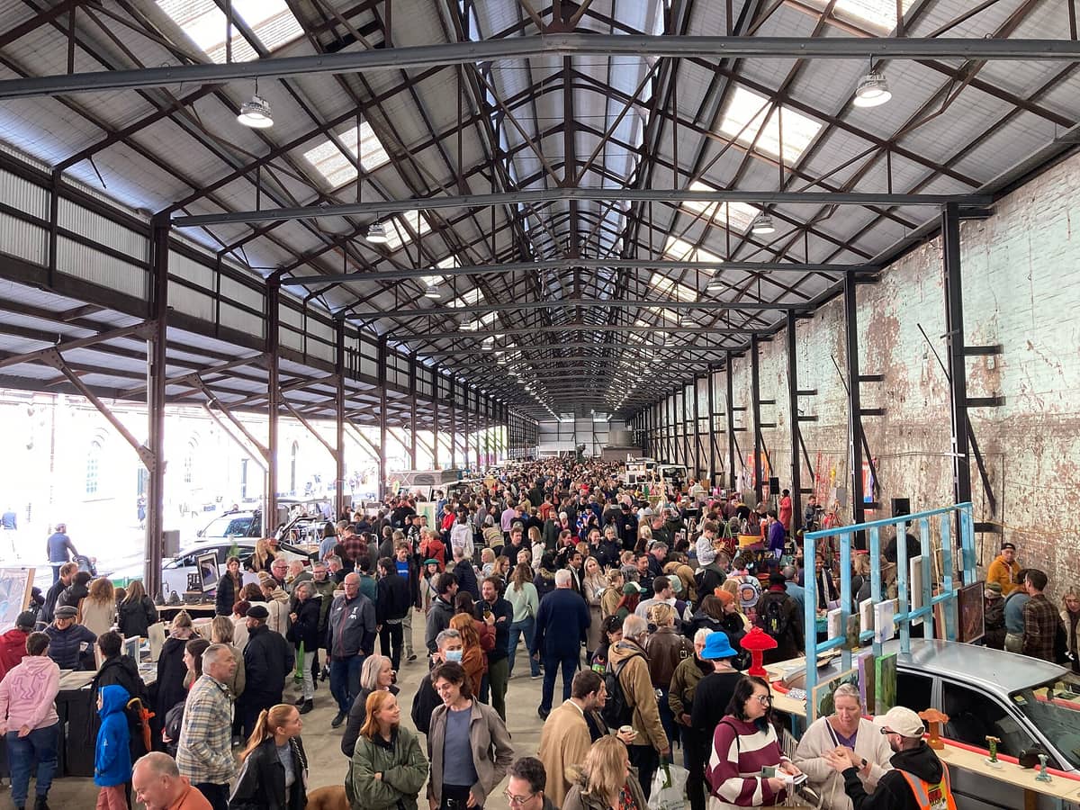 Un animado mercado interior en un gran edificio industrial, lleno de personas explorando puestos de comida y artesanía bajo un alto techo de metal.