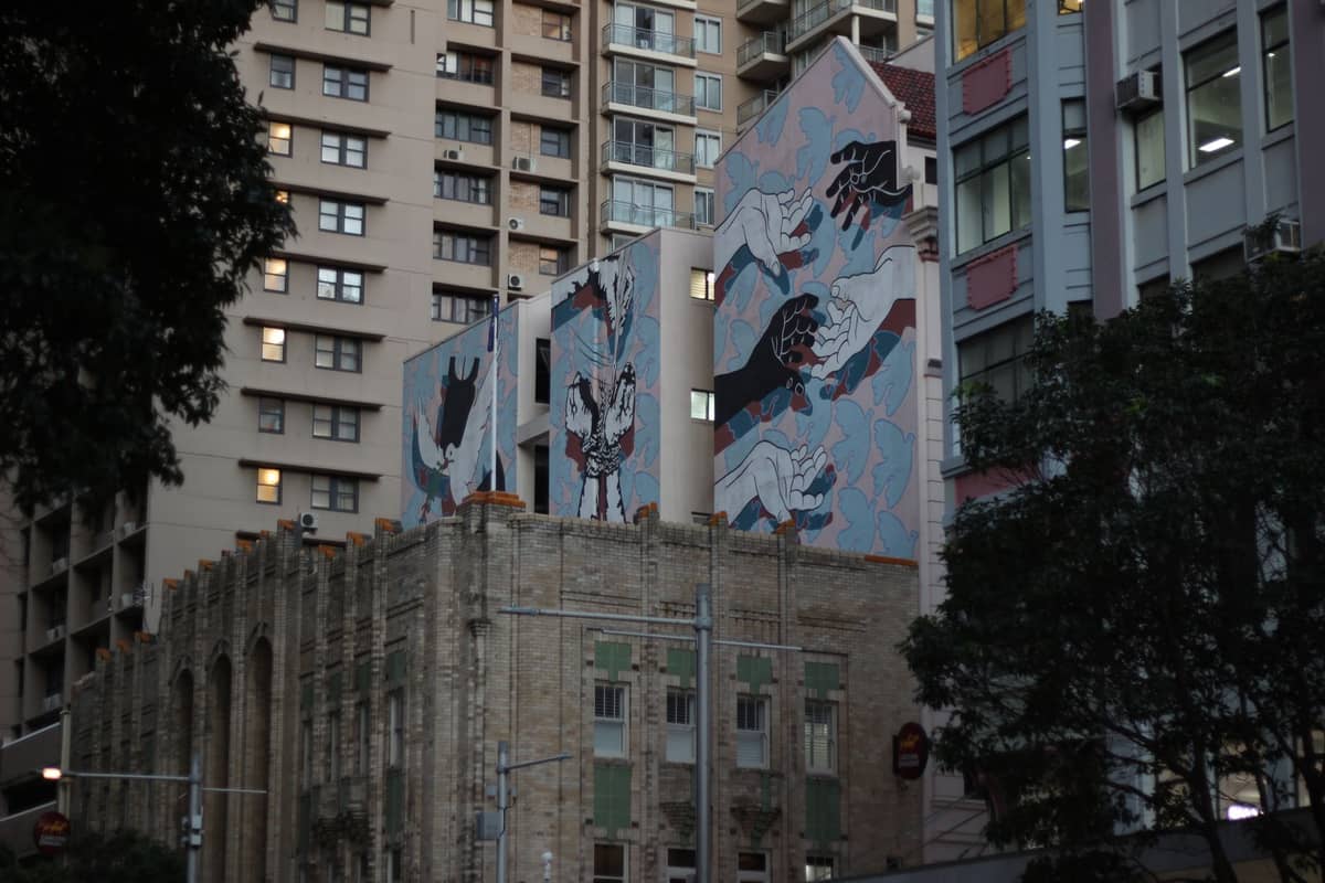 Vista de calle de tres grandes murales en edificios sobre una estructura histórica, rodeada de bloques de apartamentos modernos en Sydney.