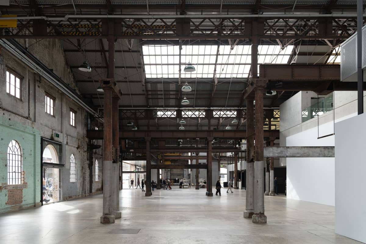 Vasto interior de un edificio industrial patrimonial con altos pilares metálicos, vigas expuestas y grandes ventanas arqueadas que dejan entrar luz natural.