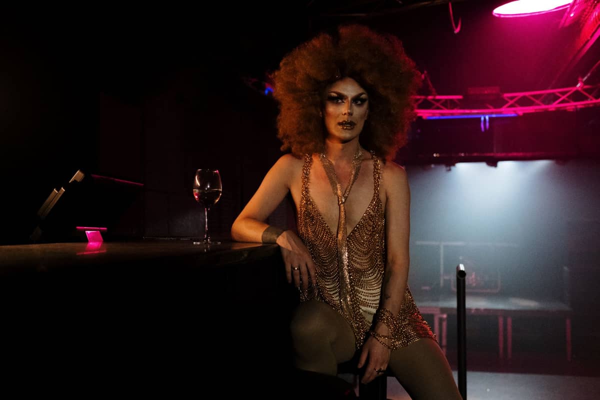 Drag-Performer in einem goldenen Kleid mit aufwendiger Frisur sitzt an einer Bar in einem Nachtclub mit Bühnenlichtern und einem Glas Wein in der Nähe.