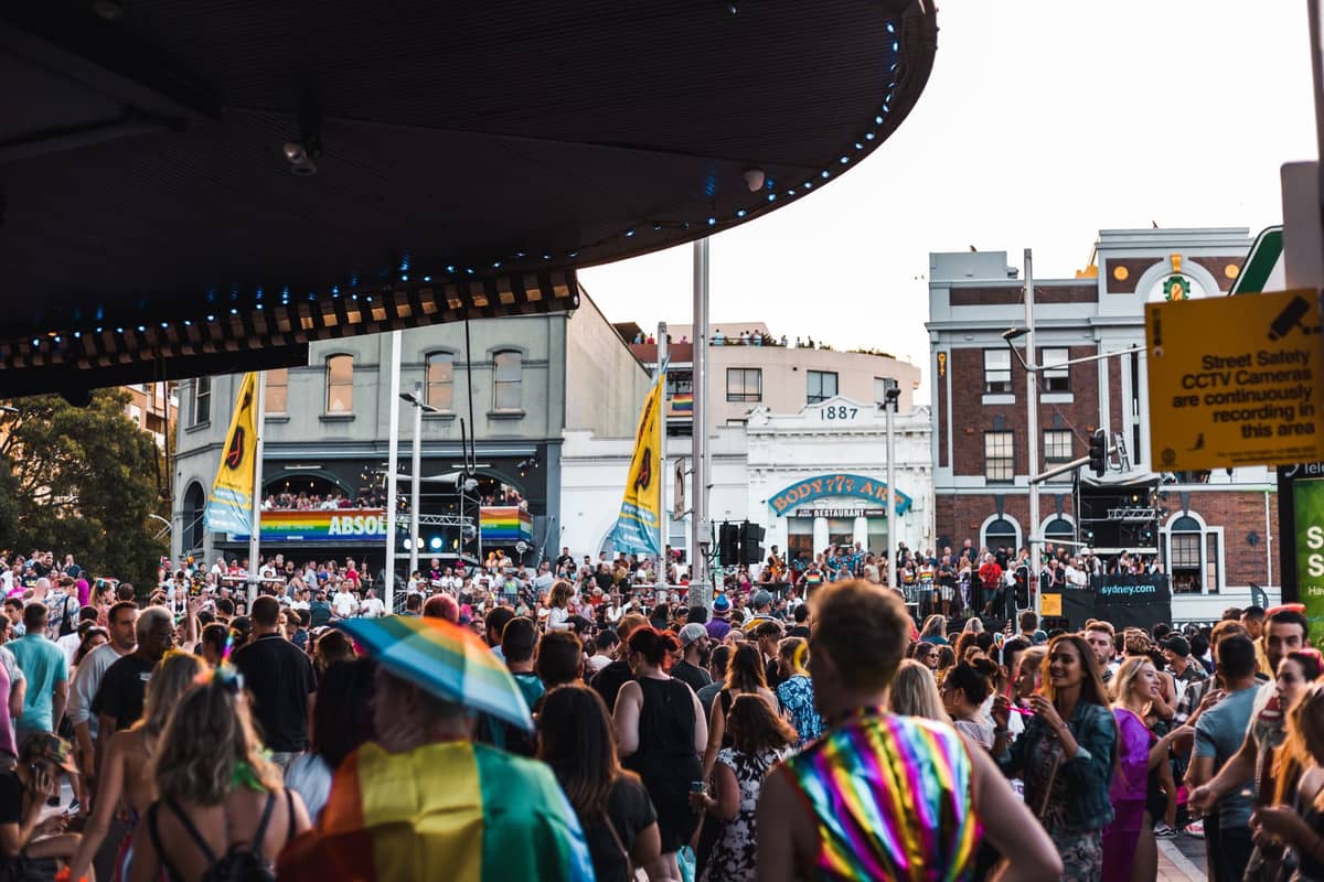 Große Menschenmengen versammeln sich während einer Mardi-Gras-Feier auf einer von klassischen Gebäuden gesäumten Stadtstraße, mit Regenbogenfahnen und festlichen Outfits.