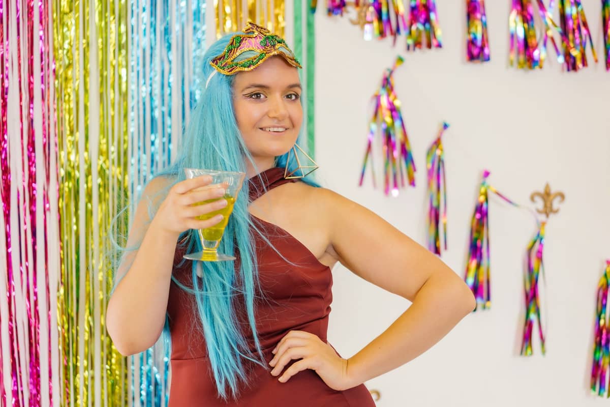 Eine Person mit blauer Perücke und festlichem Outfit hält ein Getränk in der Hand und steht vor bunten Mardi-Gras-Dekorationen in Regenbogenfarben.