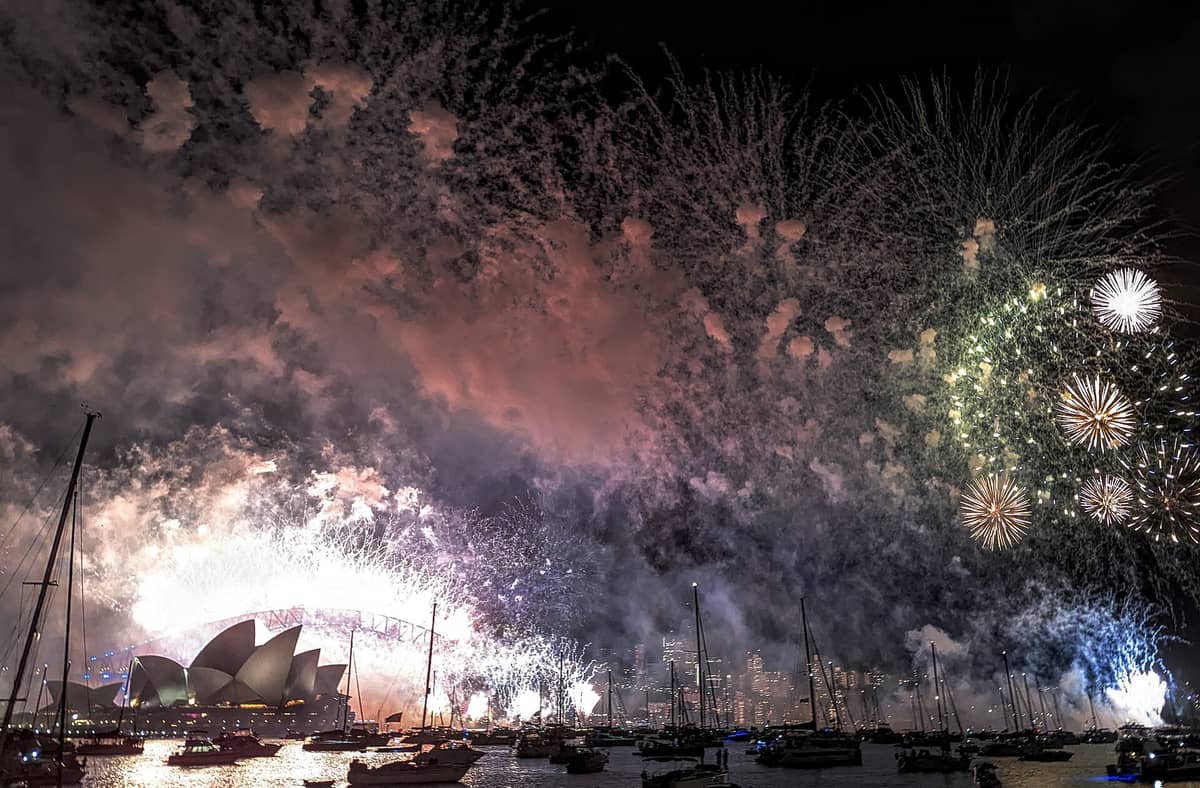 Fuegos artificiales estallan sobre Sydney Harbour con la Opera House y el Harbour Bridge visibles, botes en el agua y el horizonte de la ciudad de fondo durante la Nochevieja.