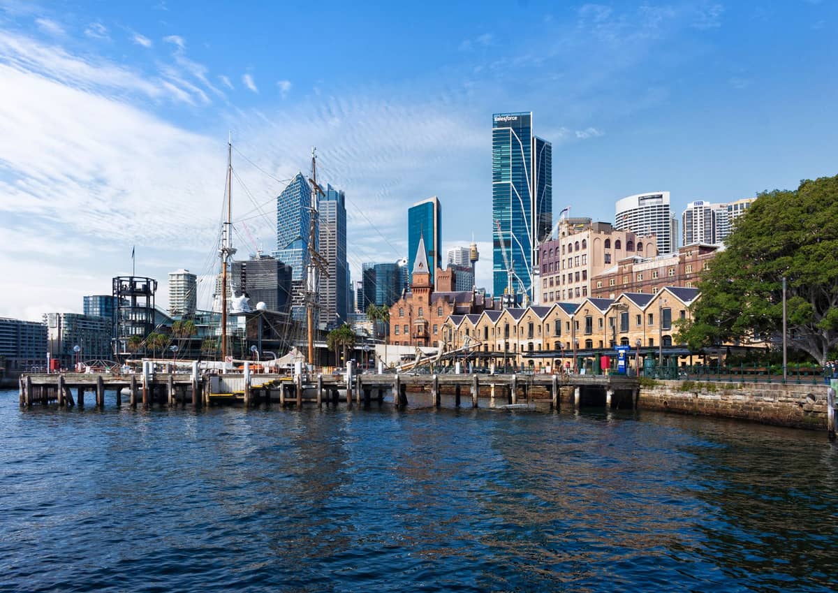 Vista del puerto de Sydney con césped en la orilla, muelles y el horizonte de la ciudad al otro lado del agua en un día despejado.