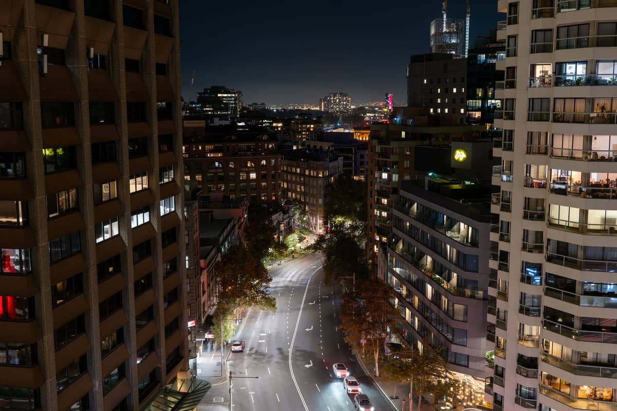 High-angle view of a lively Sydney street at night, with modern buildings, tree-lined sidewalks and light trails from passing cars.