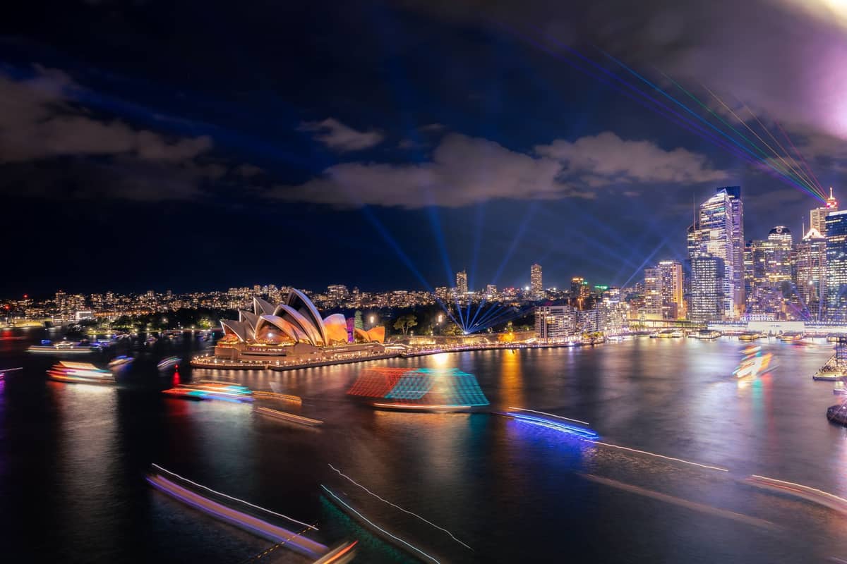 Sydney Opera House and CBD skyline lit up with colorful light projections and beams over the harbor at night during a major festival.