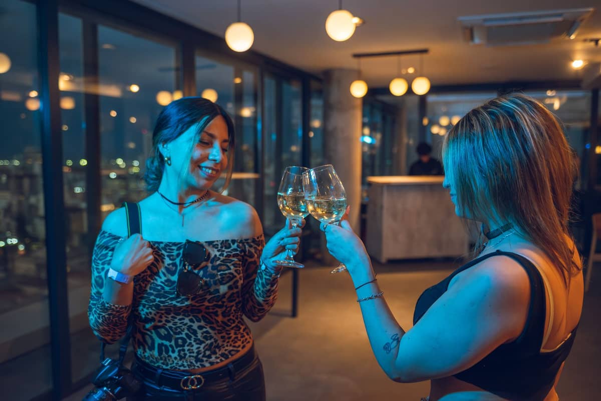 Two women inside a modern bar or lounge at night, holding wine glasses in a toast, with city lights visible through the large windows.