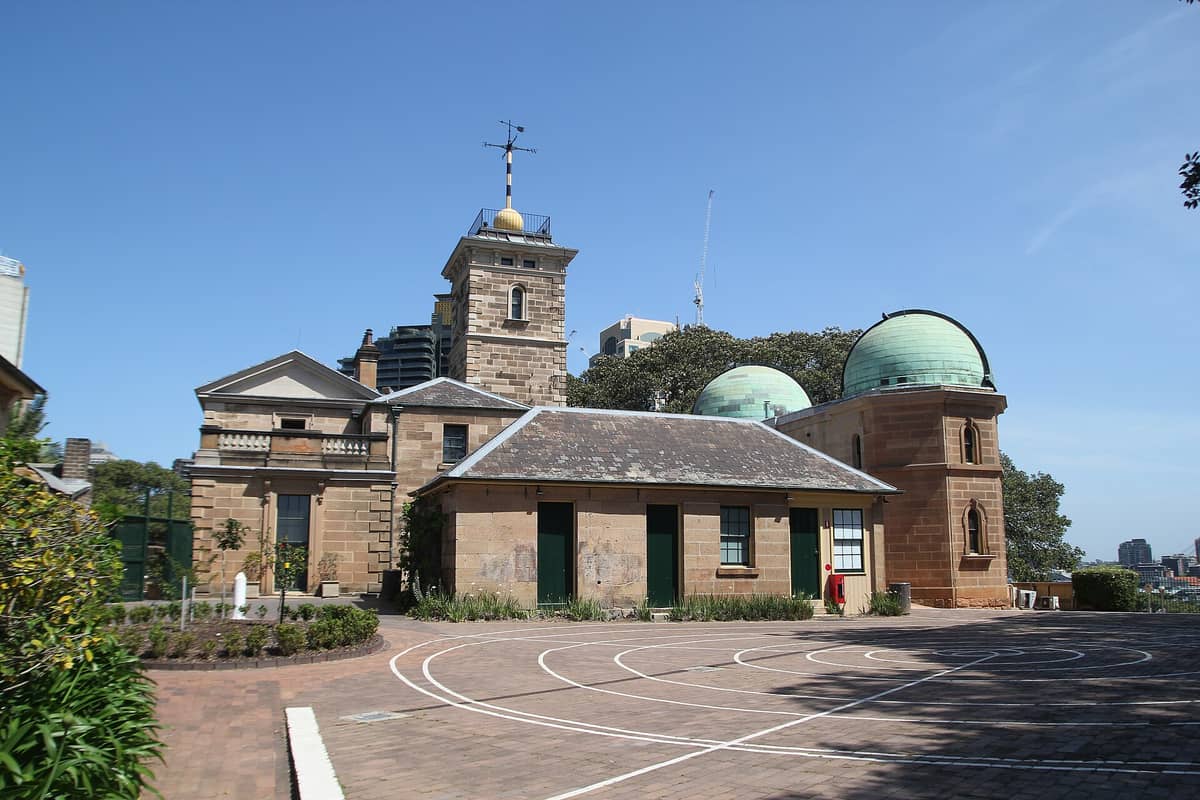 Außenansicht der Sydney Observatory mit ihrem historischen Sandsteingebäude, zwei grün gekuppelten Türmen und blauem Himmel im Hintergrund.