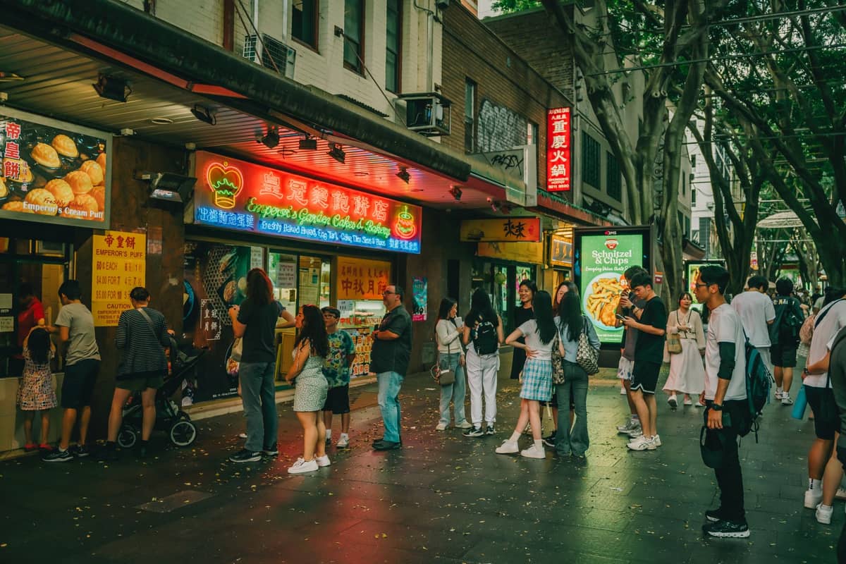 Menschen, die vor günstigen asiatischen Lokalen mit Neonreklamen in einer belebten Straße in Sydney anstehen.