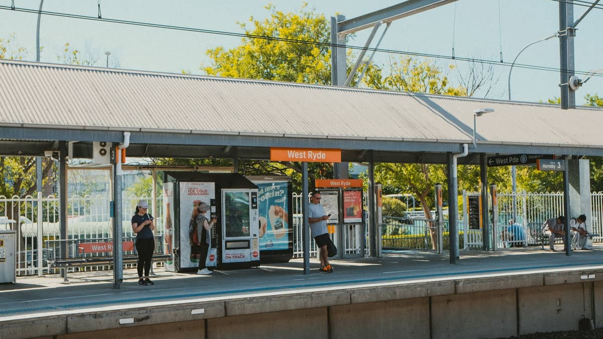 Mehrere Personen warten auf dem Bahnsteig des Bahnhofs West Ryde unter dem Unterstand, mit sichtbaren Bahnhofsschildern und Automaten.
