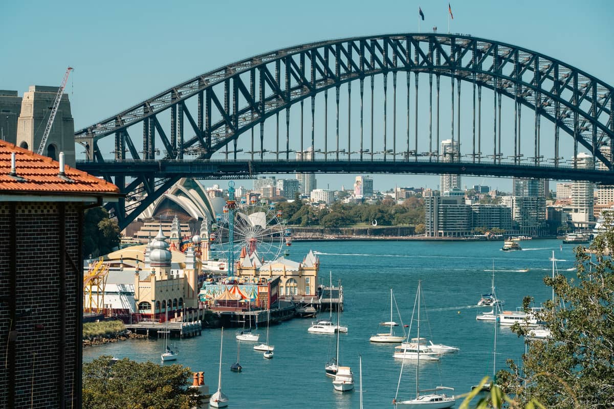Blick auf den Sydney Harbour mit dem Opera House, der Harbour Bridge und dem Luna Park, einer historischen kostenpflichtigen Attraktion, von der anderen Seite des Wassers mit Booten.