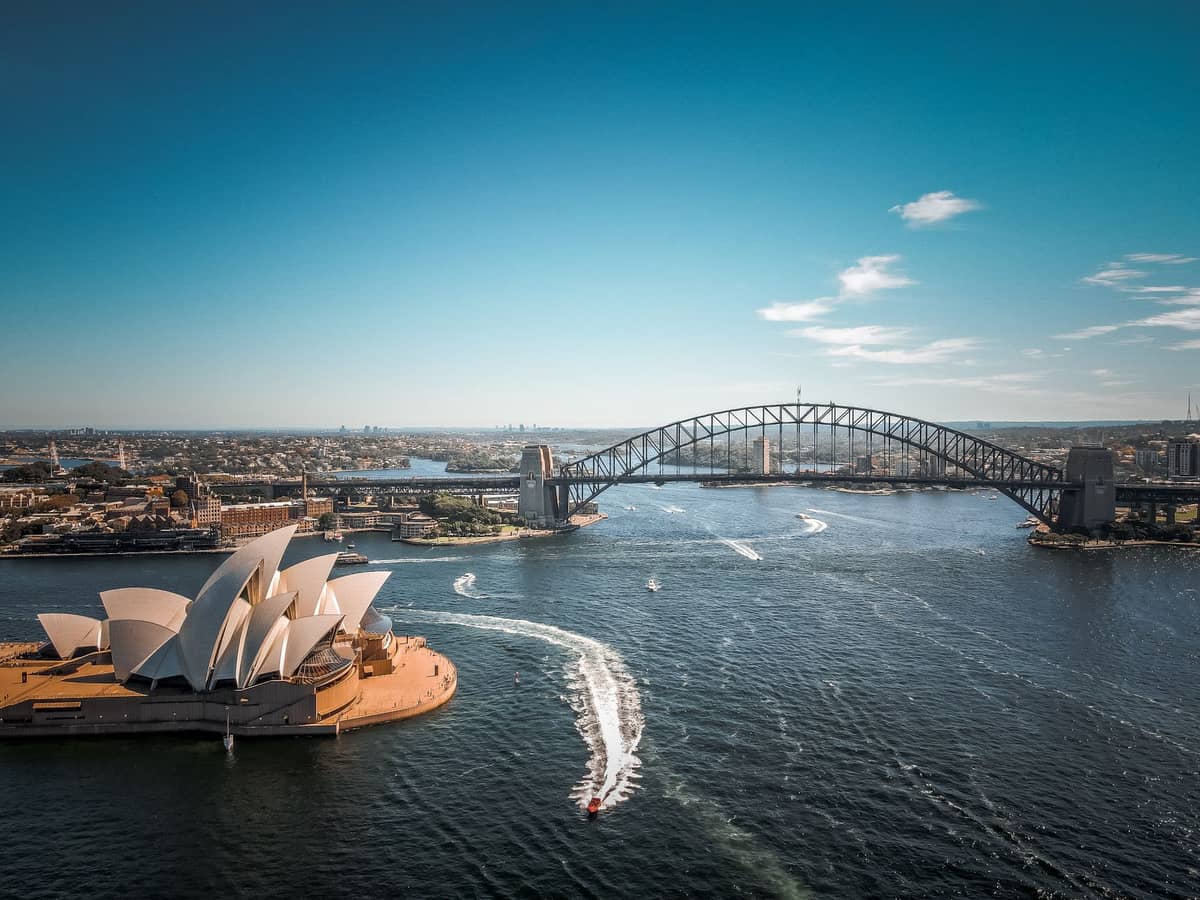 Szerokie ujęcie z lotu ptaka na Sydney Harbour z Operą w pierwszym planie i Mostem Harbour Bridge w tle pod bezchmurnym, błękitnym niebem.
