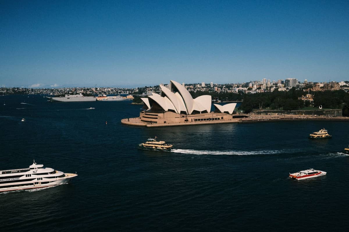 Panoramiczny widok na Sydney Opera House sfotografowany z drugiego brzegu portu, z kilkoma promami i łodziami na pierwszym planie i bezchmurnym błękitnym niebem.