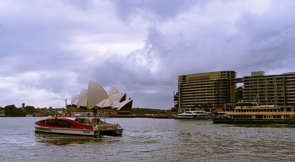 Sydney Harbour with several ferries on the water, Sydney Opera House and city skyline in background under a cloudy sky