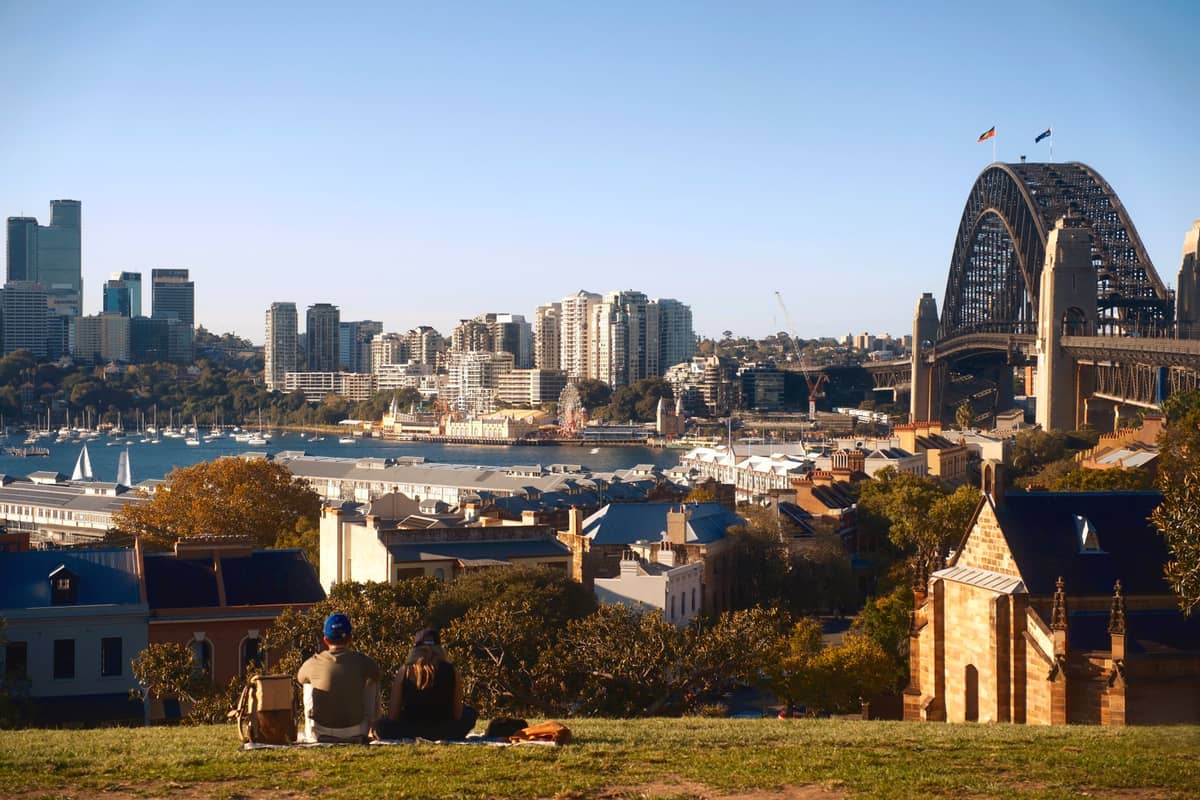 Wide view of Sydney Harbour with families sitting on a grassy hill overlooking the city, harbour, and Sydney Harbour Bridge under clear blue skies.