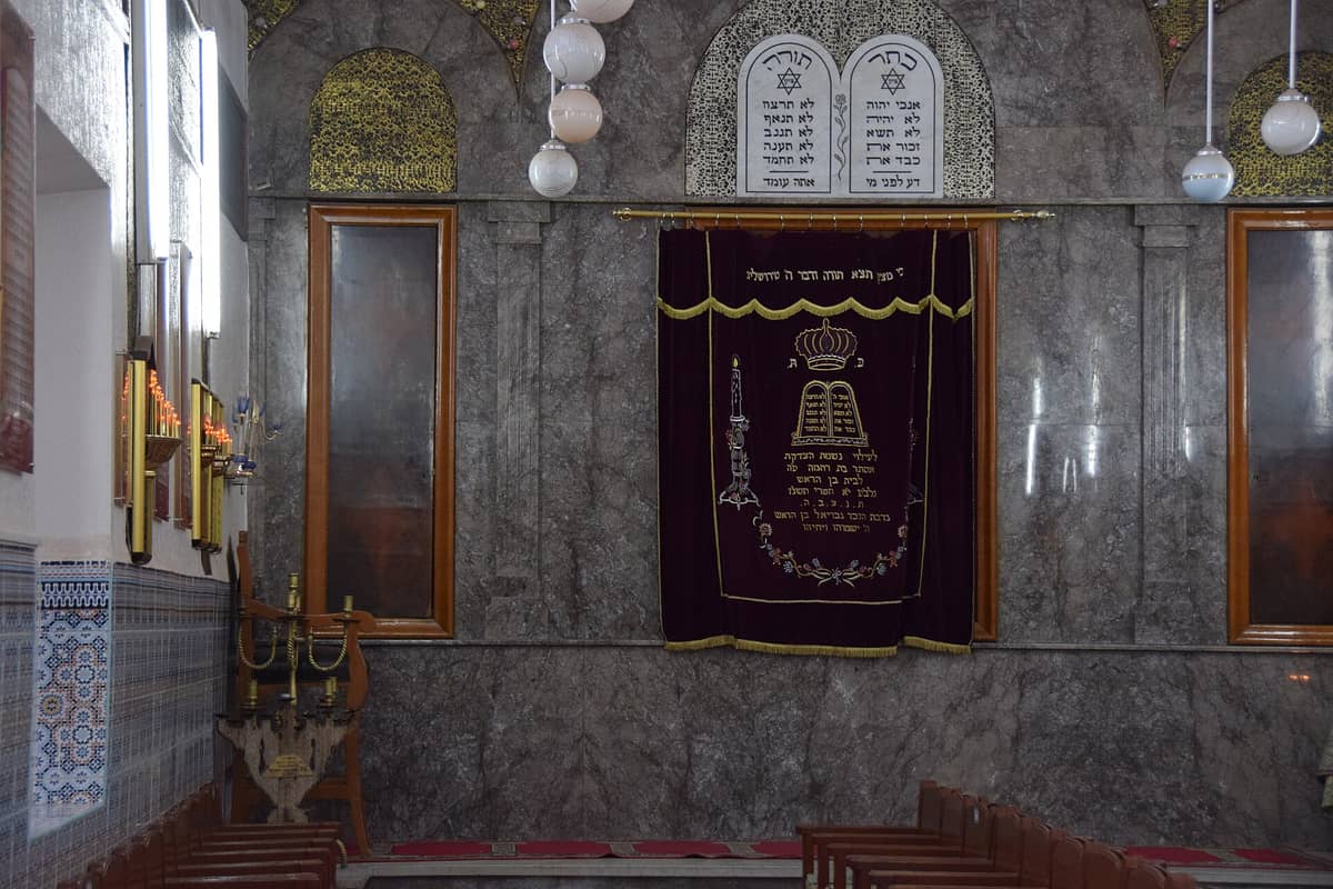 บรรยากาศภายในโบสถ์ Slat Al Azama เมืองมาราเกช ม้านั่งอธิษฐาน หีบ Torah ผ้าม่านกำมะหยี่ และจารึกภาษาฮีบรูบนผนังหินอ่อน