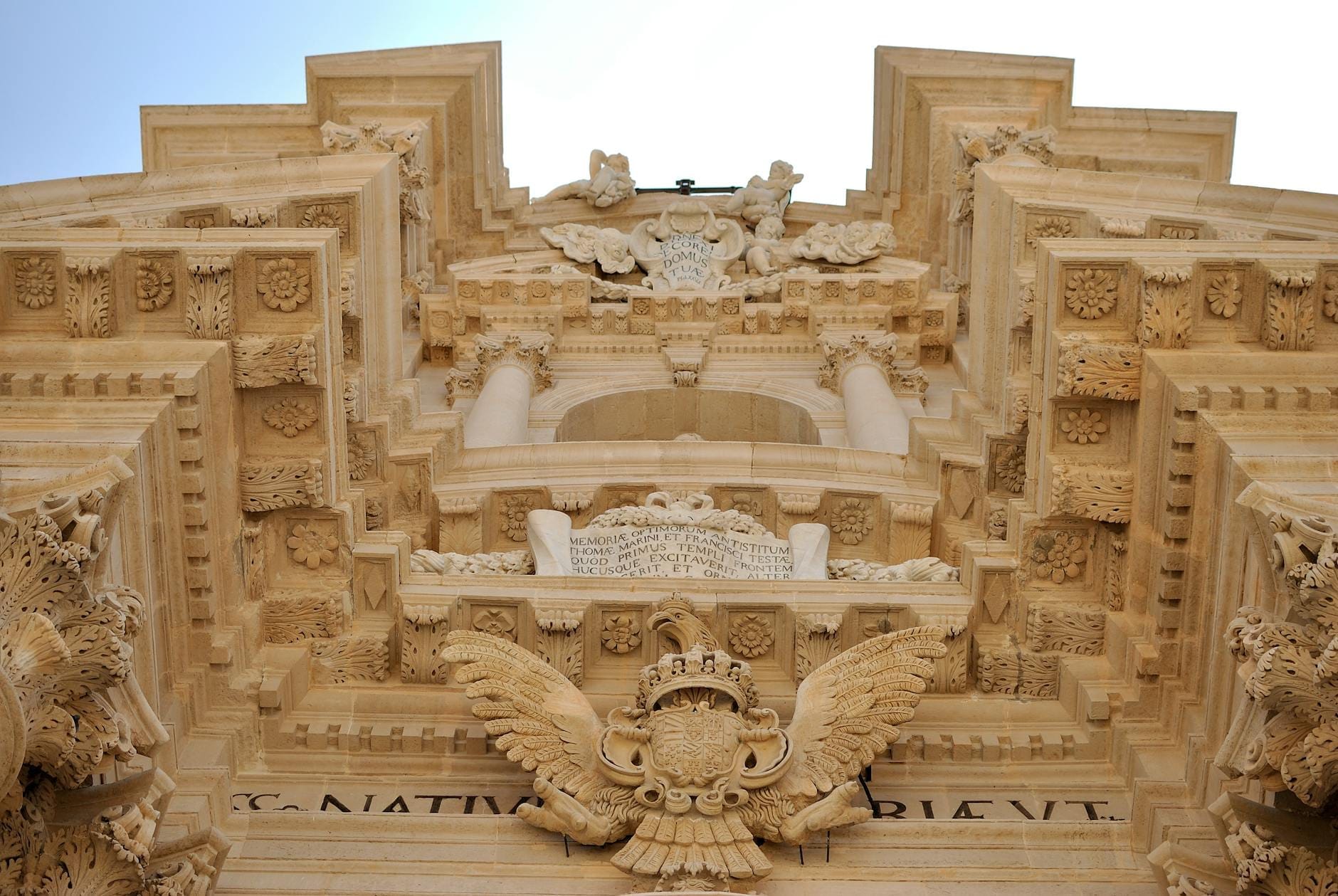 Dramatische Untersicht der reich verzierten Barockfassade der Kathedrale von Syrakus mit fein gearbeiteten Steinreliefs und klassischen Säulen vor einem klaren Himmel.