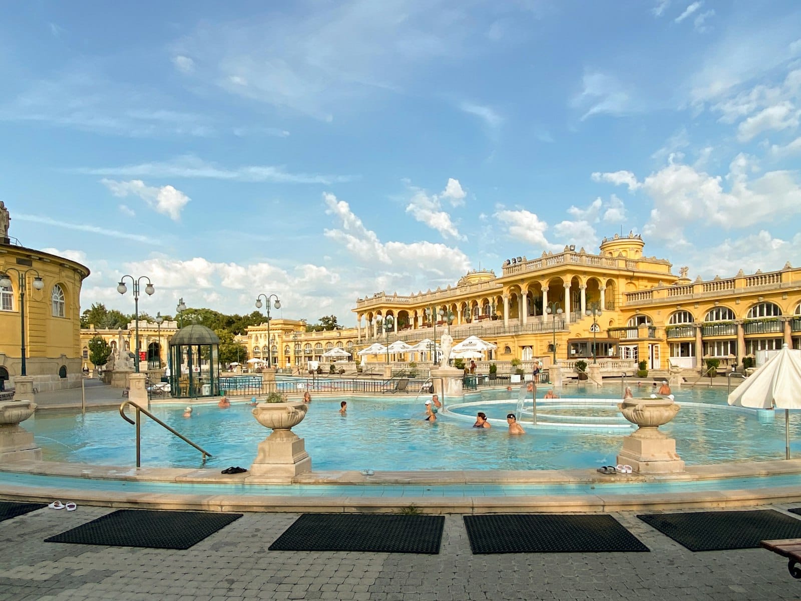 Bassin thermal extérieur des thermes Széchenyi avec des baigneurs se détendant, encadré par l'architecture néo-baroque du palais jaune sous un ciel bleu lumineux.