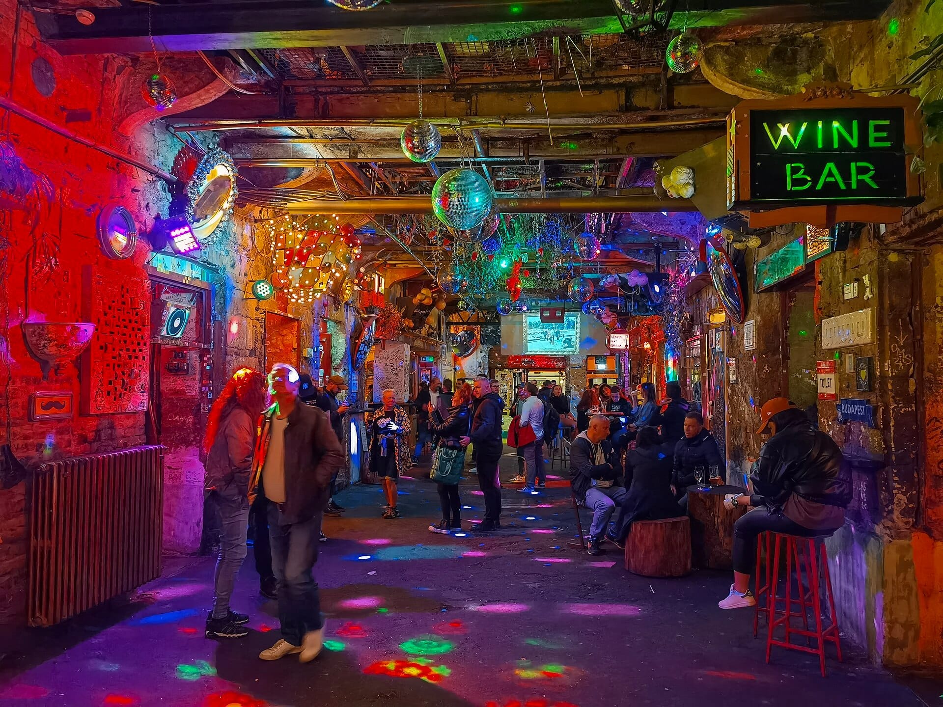 Interior colorido del bar de ruinas Szimpla Kert en Budapest, con paredes de ladrillo expuesto, letreros de neón, bolas de discoteca y grupos de personas socializando.