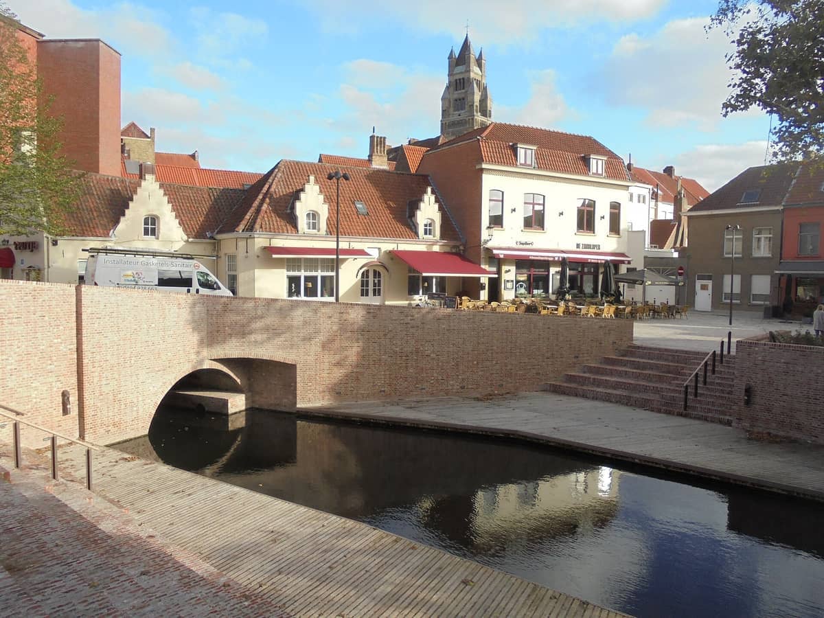 View of 't Zand in Bruges featuring a brick canal bridge, terraced cafes, historic gabled buildings, and the Concertgebouw spire under a partly cloudy sky.