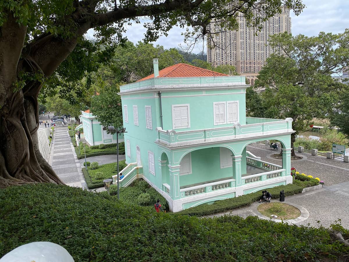 Blick auf die hellgrünen portugiesischen Kolonialvillen des Taipa Houses-Museums in Macau, mit zwei der historischen Uferhäuser und gepflegten Wegen zwischen Bäumen und Gartenanlagen.