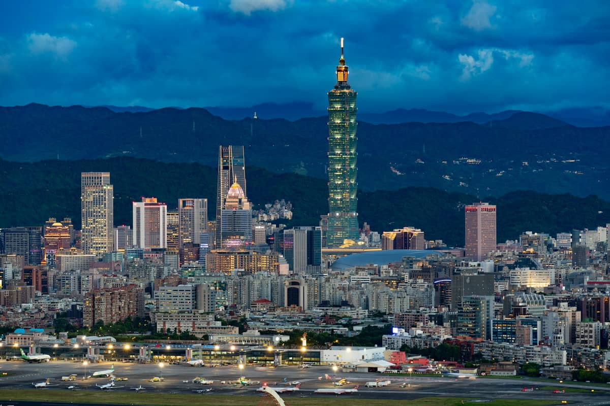 Panoramablick auf die Skyline von Taipei in der Abenddämmerung, mit den Landebahnen des Flughafens Songshan im Vordergrund und dem Taipei 101 im Hintergrund.