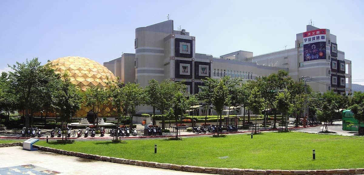 Außenansicht des Taipei Astronomical Museum in Shilin mit seiner ikonischen goldenen Kuppel, modernen Gebäuden, grünen Rasenflächen und geparkten Rollern unter sonnigem Himmel.