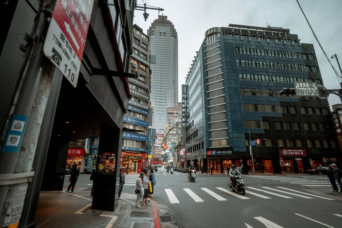 Stadtstraße in Taipei, gesäumt von mittelhohen Gebäuden, Geschäften und Restaurants, belebt von Fußgängern, Motorrädern und modernem Stadtleben.