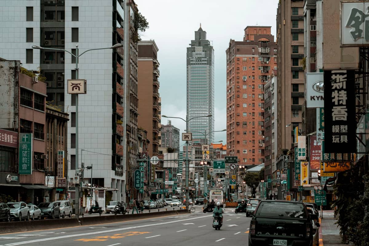 Straßenansicht der Innenstadt von Taipei mit lebhaftem Verkehr, Scootern, Schildern auf Chinesisch und dem markanten Shin Kong Life Tower im Hintergrund.