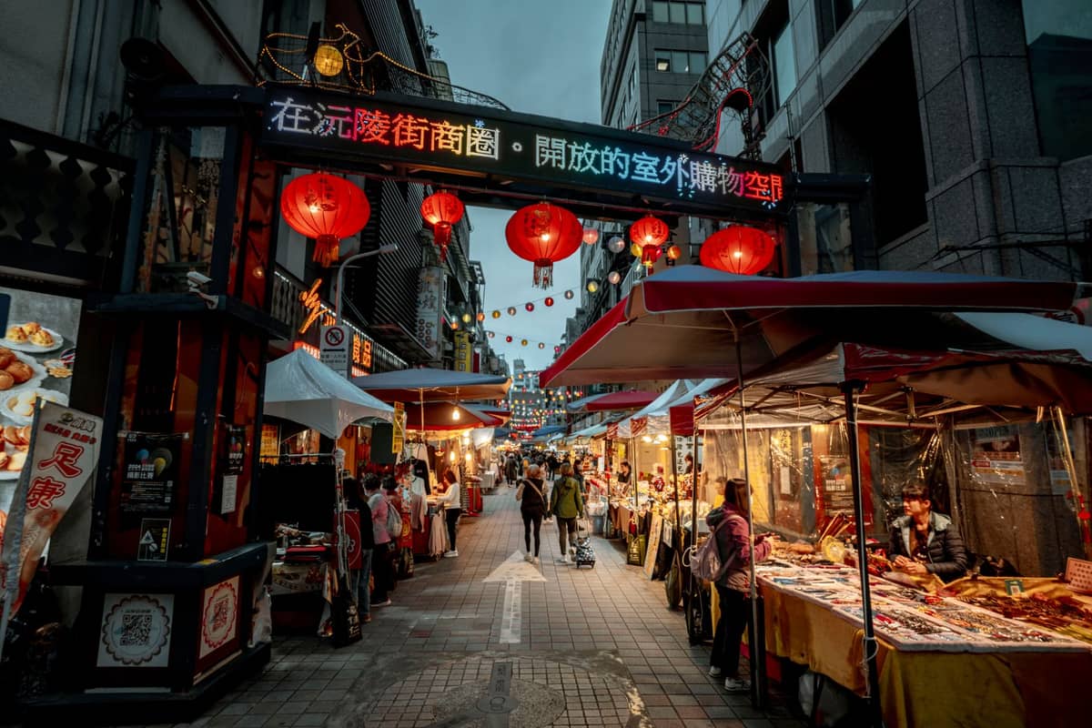 Abendlicher Blick auf einen belebten Straßenmarkt in Taipei mit Essensständen, roten Laternen und Menschen, die unter einem dekorativen Tor spazieren.