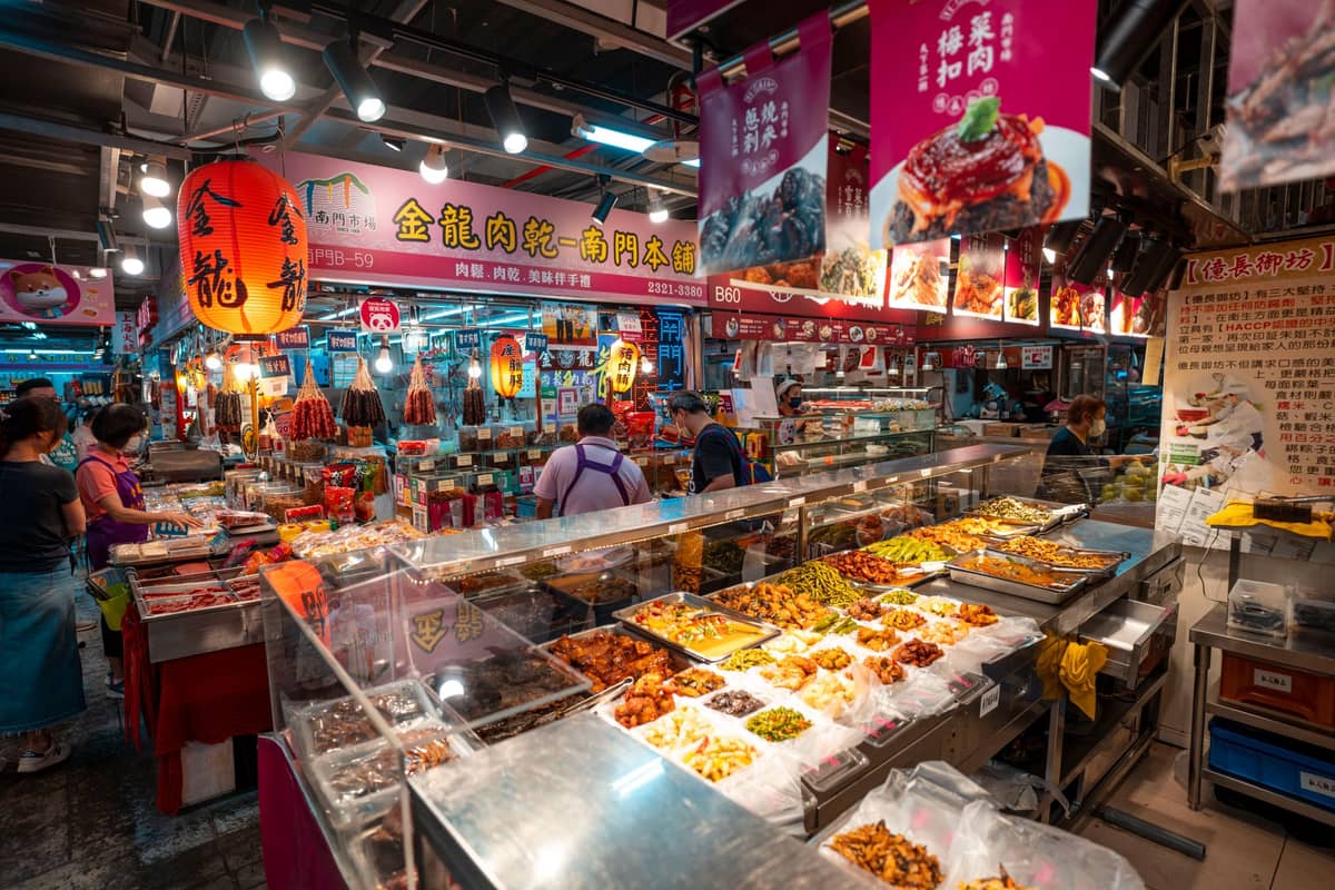 Ein lebhafter Lebensmittelmarkt in Taipeh mit verschiedenen taiwanesischen Gerichten in Schalen, beleuchteten Schildern auf Chinesisch und Menschen, die durch die Stände schlendern.