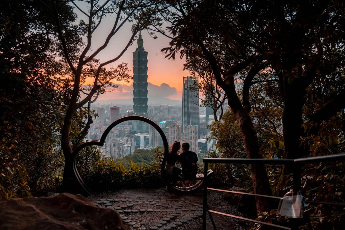 Aussicht vom Elephant-Mountain-Trail in Taipeh bei Sonnenuntergang – Blick auf den Taipei 101 durch Bäume, Wanderer sitzen am Aussichtspunkt.