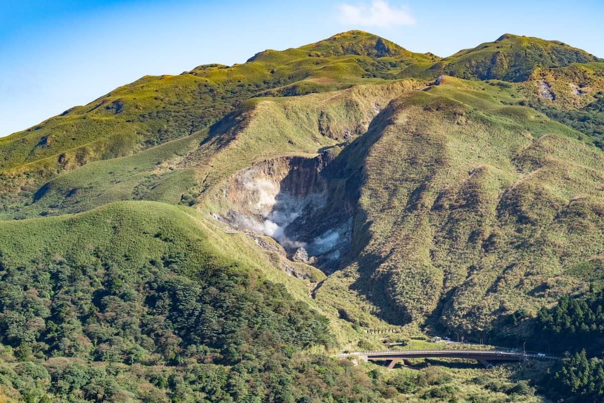 Landschaftsansicht des Vulkankraters von Yangmingshan, mit aufsteigendem Dampf aus den Fumarolen, umgeben von üppig grünen Hügeln unter blauem Himmel.