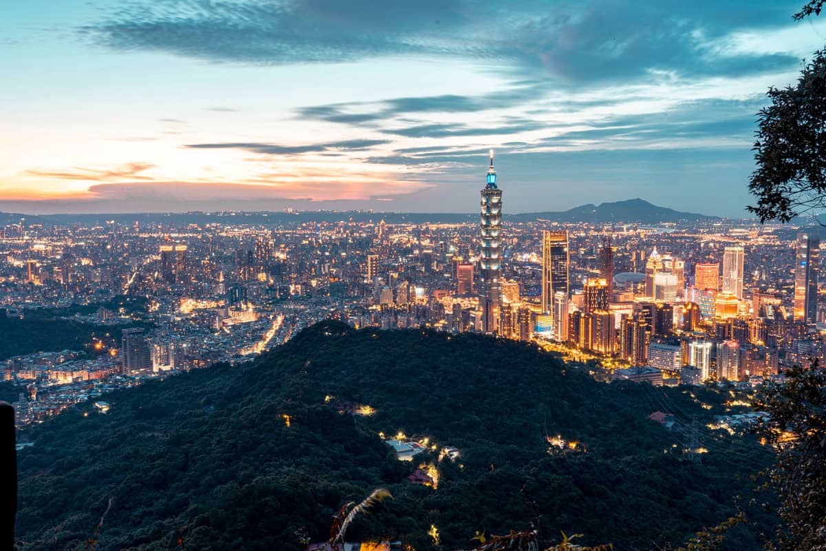 Weite Landschaftsansicht von einem städtischen Wanderweg mit Blick auf die Skyline von Taipei, Taipei 101 und bewaldete Hügel bei Sonnenuntergang.
