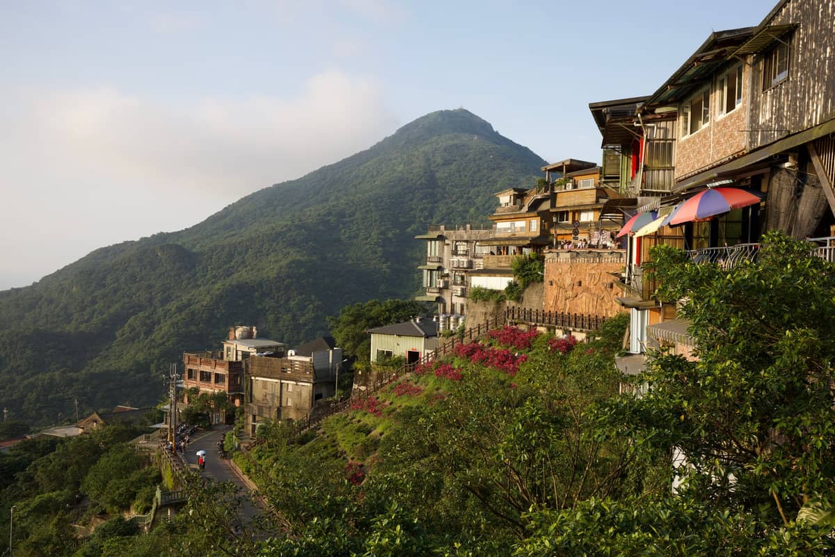 Bergdorf mit Gebäuden am Hang, Grünflächen und einem Berggipfel im Hintergrund in der Nähe von Taipei, im Licht des späten Nachmittags.