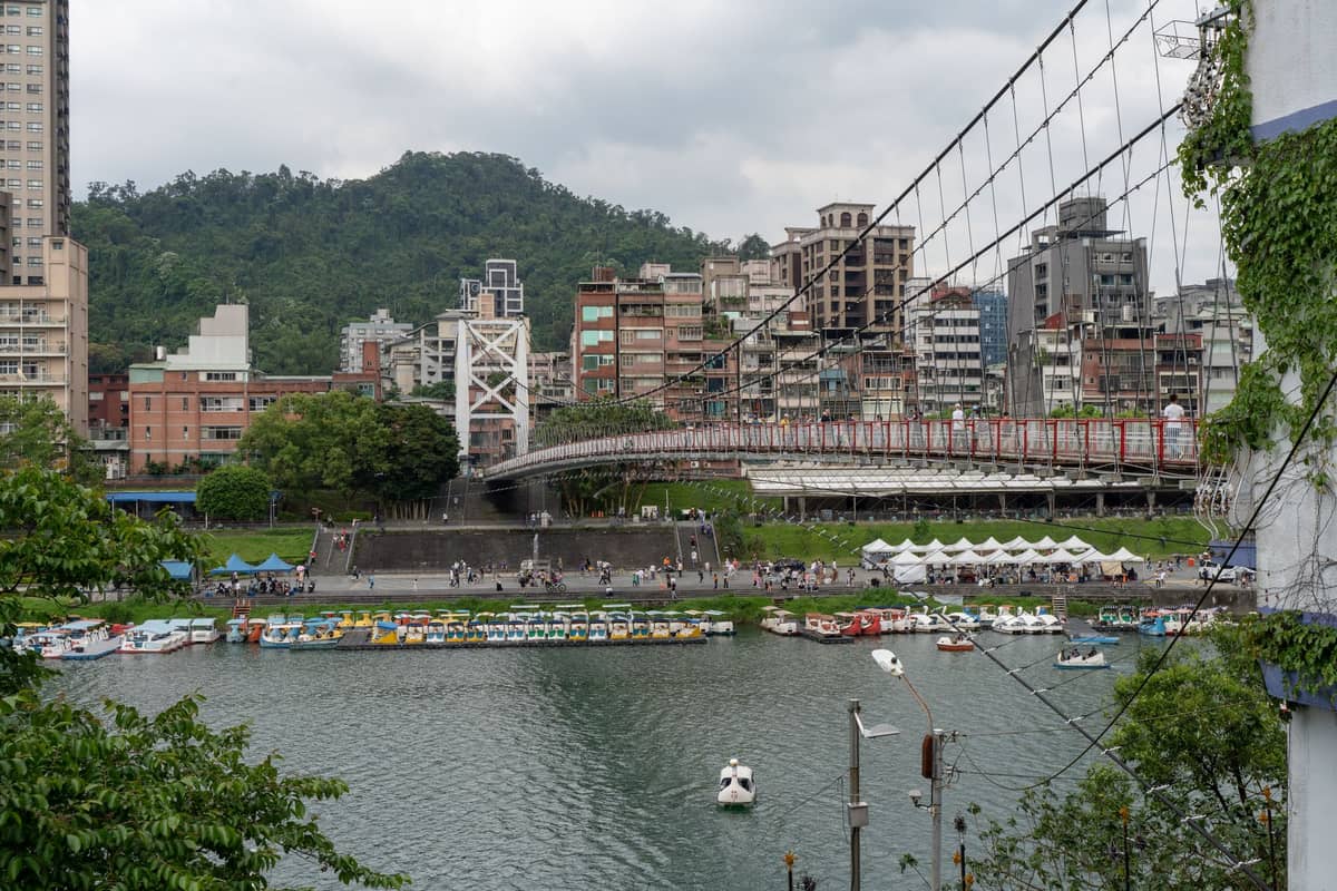 Hängebrücke und Flussuferszene mit Booten, Stadtgebäuden und grünen Hügeln, die an Taipeis zugängliche städtische Ausflugsziele nahe den Thermalquellenbezirken erinnern.