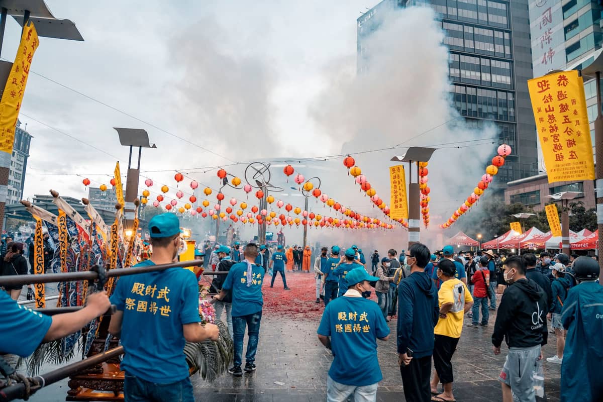 Menschenmenge in blauen Shirts bei einem lebhaften Straßenfest mit Laternen, Rauch, Bannern und Hochhäusern von Taipei im Hintergrund.
