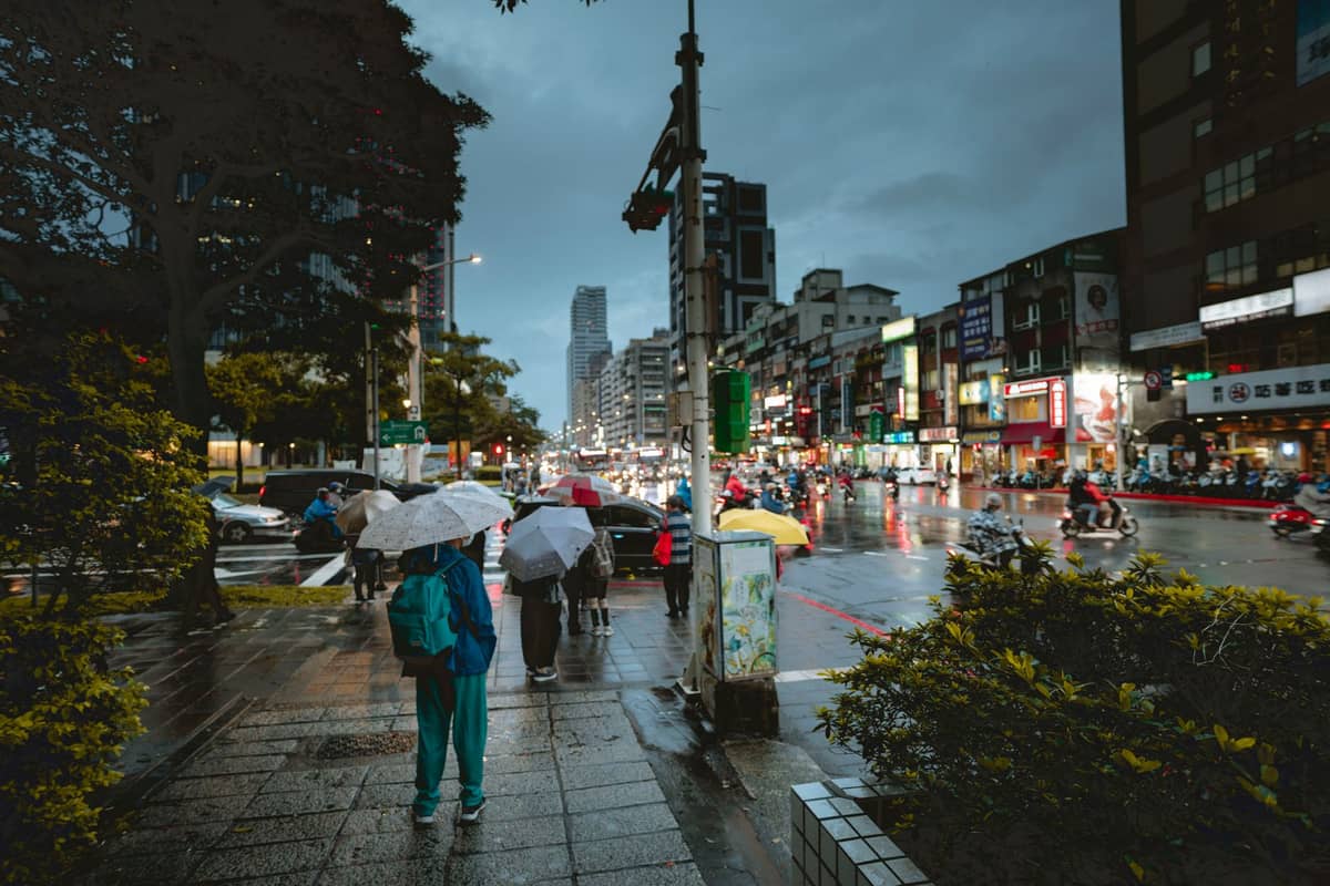 Menschen mit Regenschirmen auf einem nassen Stadtbürgersteig in Taipei, mit glänzenden regengetränkten Straßen und grauem Frühlingshimmel.