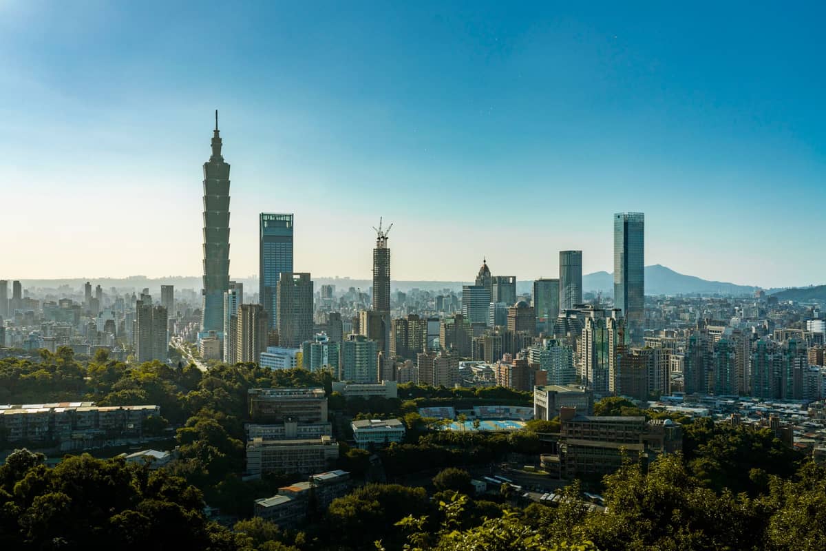 Tagsüber aufgenommene Panoramaansicht der Skyline von Taipei mit dem Taipei 101-Turm und modernen Wolkenkratzern, aufgenommen von einem Hügel mit üppigem Grün im Vordergrund.