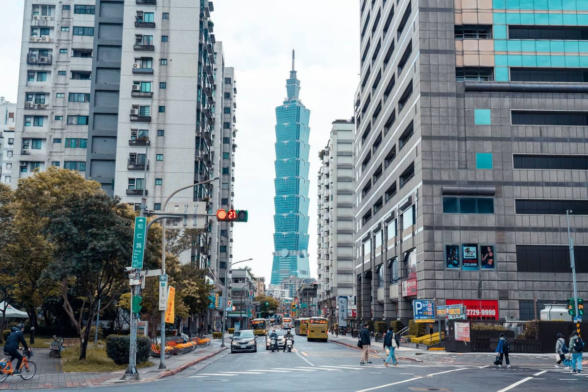 Der Taipei 101 ragt markant zwischen Stadtstraßen und Gebäuden empor, im Vordergrund Menschen und Verkehr unter einem bewölkten Himmel.