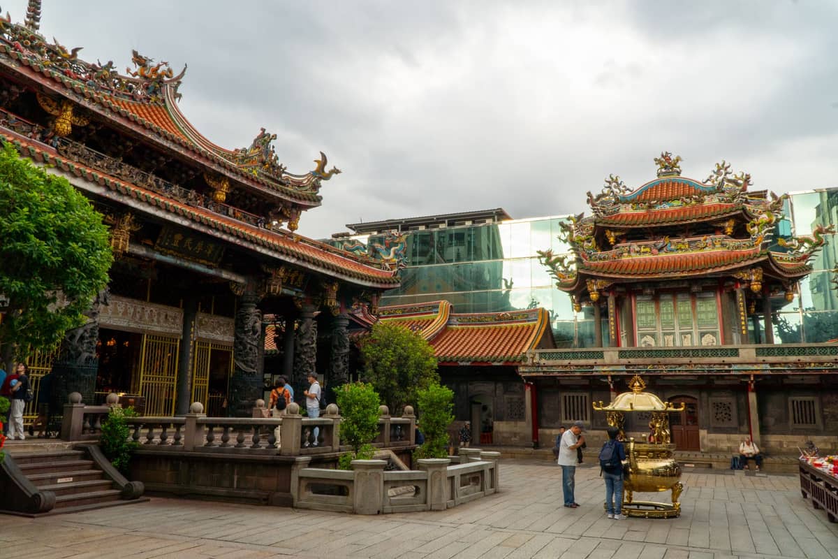 Weitwinkelansicht des Innenhofs des Lungshan-Tempels in Taipei mit kunstvollen Tempelstrukturen, Besuchern und modernen Glasgebäuden im Hintergrund.