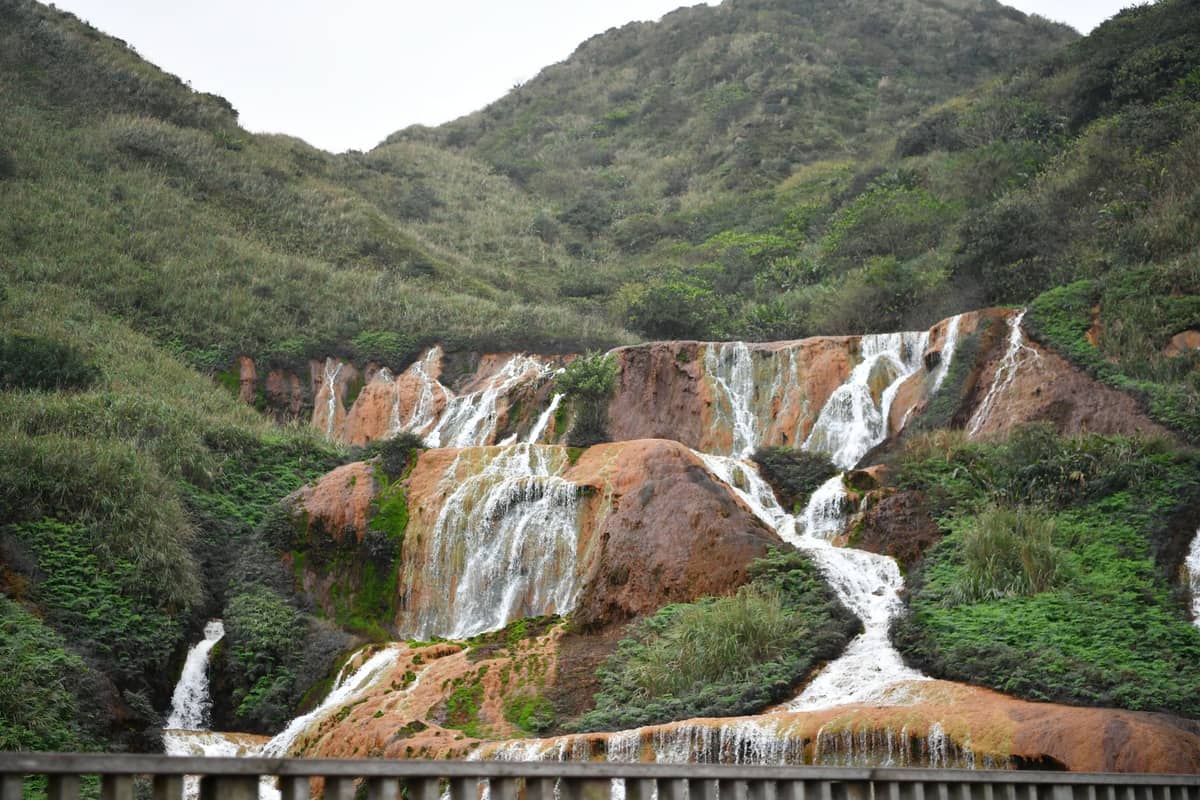 Goldener Wasserfall in Jinguashi, Taiwan, mit leuchtend orangefarbenen Felsen, kaskadierendem Wasser und üppigen grünen Hügeln im Hintergrund.