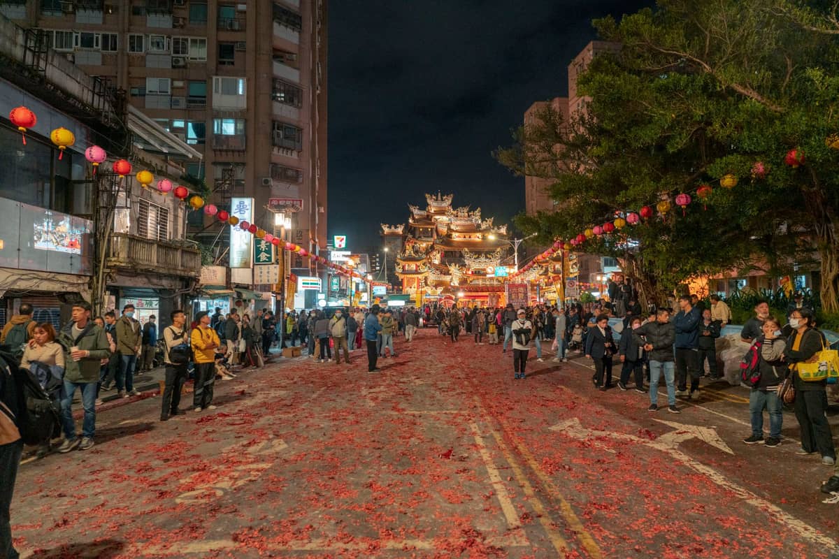 Abendliche Straßenszene in Taipei mit einer Menschenmenge, festlichen Laternen über den Köpfen und einem beleuchteten Tempel im Hintergrund.