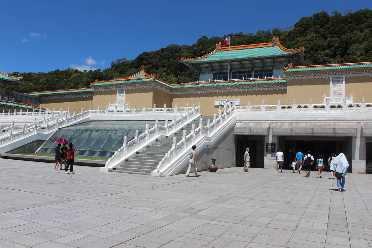 Weitwinkelbild des Eingangs des National Palace Museum mit Treppen, traditionellen chinesischen Gebäuden und Besuchern an einem sonnigen Tag.