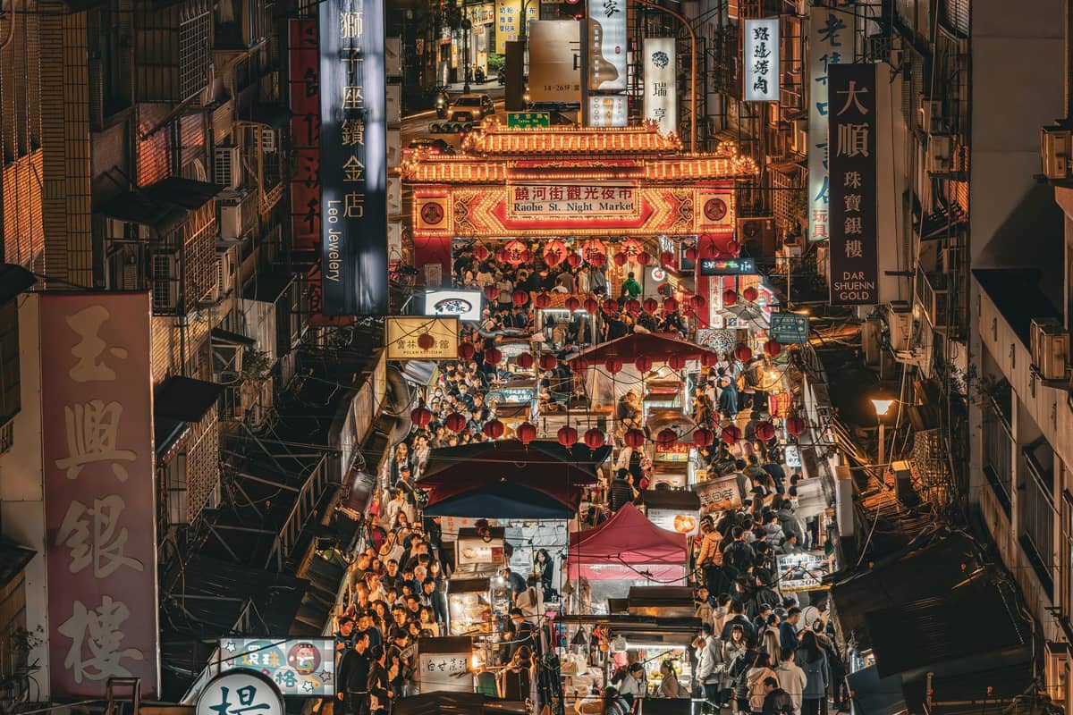 Luftaufnahme eines belebten Nachtmarkts in Taipei, mit zahlreichen Essensständen, Menschenmassen und dem beleuchteten Eingangstor des Raohe Street Night Market.