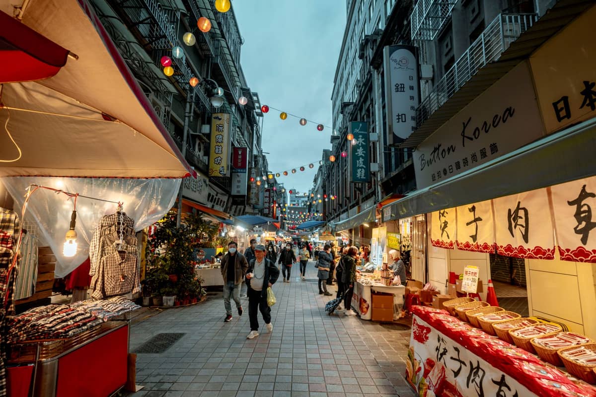 Belebte Straße in Taipei am Abend mit traditionellen Ladenfront, hängenden Laternen, Essensständen und Menschen, die unter Marktlichtern spazieren.