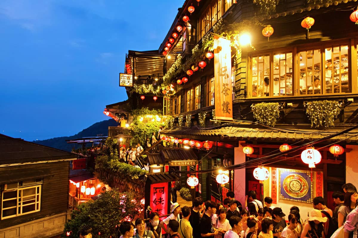 Menschenmassen versammeln sich nachts neben einem laternenbeleuchteten Teehaus in Jiufen, mit leuchtend roten Lichtern und Hangblicken unter einem blauen Abendhimmel.