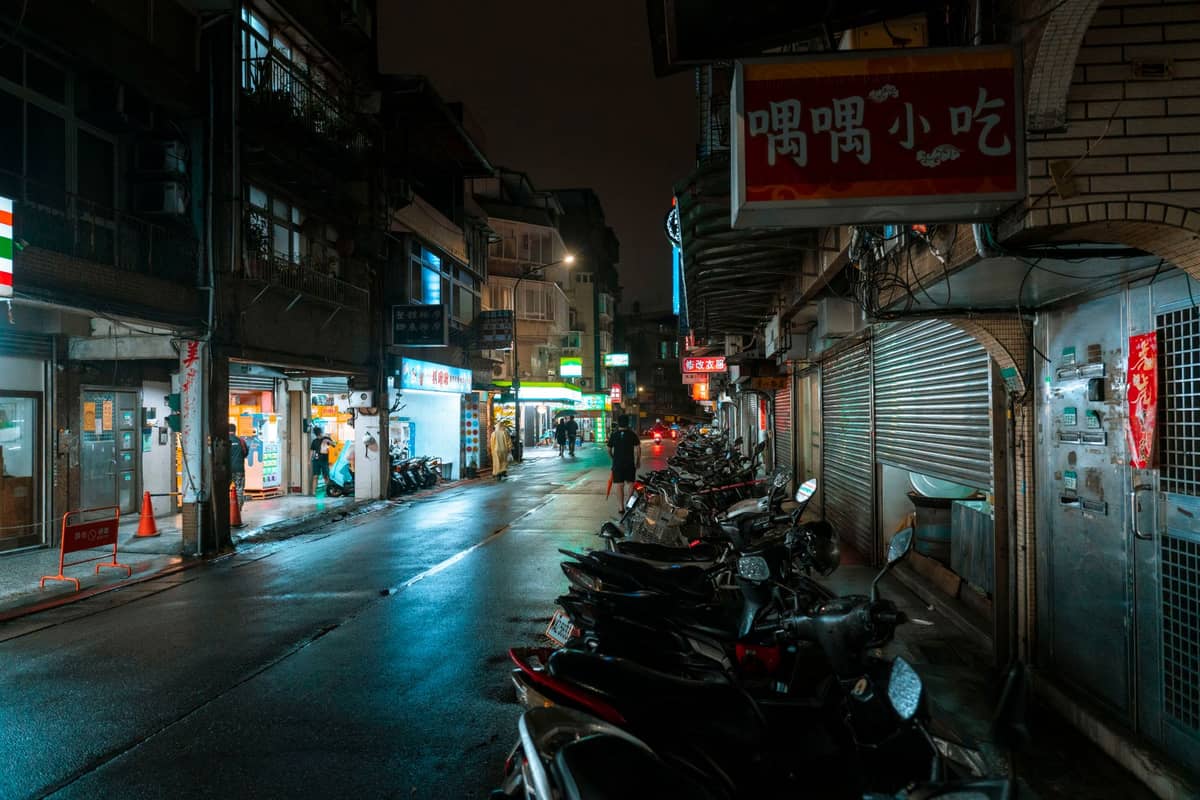 Enge Straße in Taipei bei Nacht, gesäumt von Rollern und kleinen Restaurants mit hellen Schildern, die die Atmosphäre in der Nähe von Nachtmärkten heraufbeschwören.