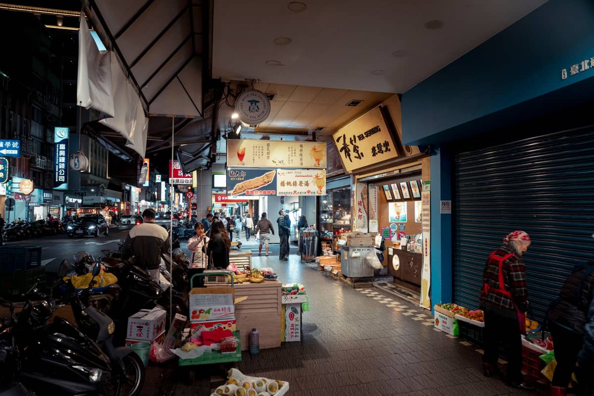 Eine ruhige nächtliche Marktszene in einem Taipei-Viertel mit Einheimischen, die an kleinen Ständen und Obstverkäufern entlang des Bürgersteigs einkaufen.